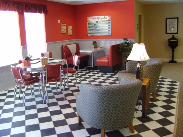 A retro-style dining area with black and white checkered flooring, red walls, and a mix of seating including red vinyl booths, red chairs around a round table, and two patterned armchairs with a small wooden table and lamp between them. A menu board and framed pictures hang on the walls, and large windows with red curtains let in natural light.