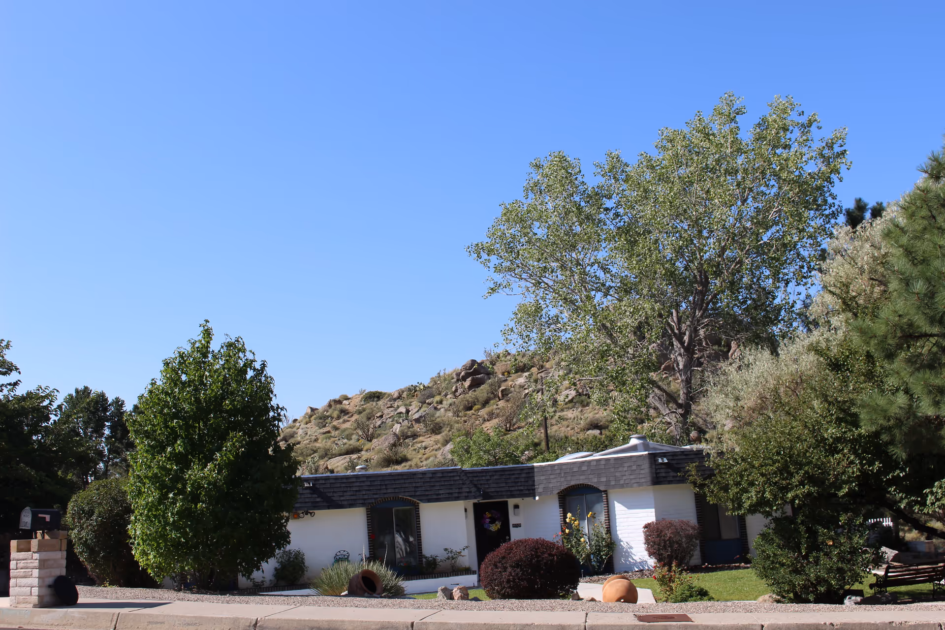 Single-story white residential building with a dark shingled roof, surrounded by various green trees and bushes, with a rocky hill in the background under a clear blue sky.