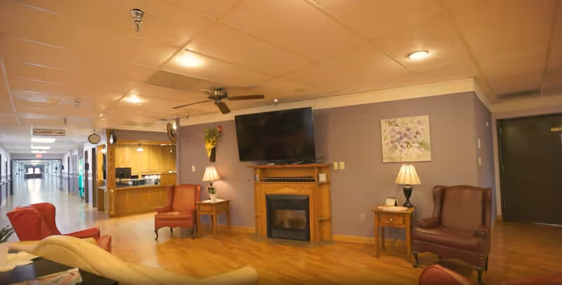 A cozy common area in a senior living facility with wooden flooring, a wall-mounted flat screen TV above a wooden fireplace, two armchairs with side tables and lamps, and a hallway leading to other rooms. The walls are painted light purple and decorated with a floral painting and a wall-mounted flower arrangement.
