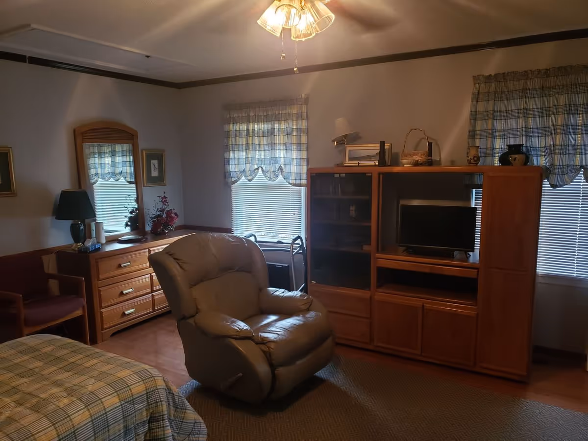 Cozy assisted-living bedroom with a recliner, wooden entertainment cabinet and TV, dresser, and a partially visible bed.