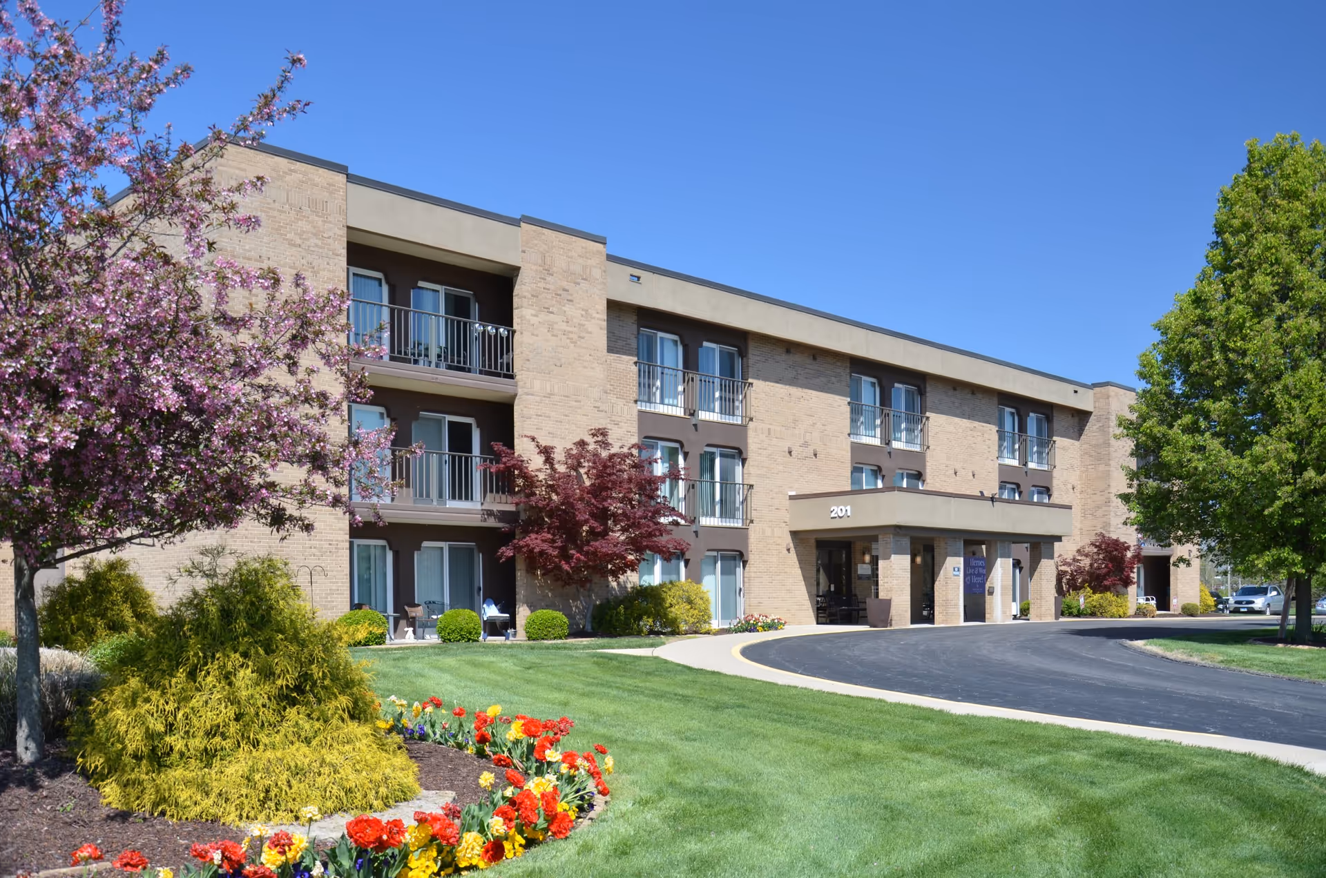 Exterior view of a three-story senior living facility building with balconies, surrounded by well-maintained landscaping including green grass, colorful flowers, and trees under a clear blue sky.