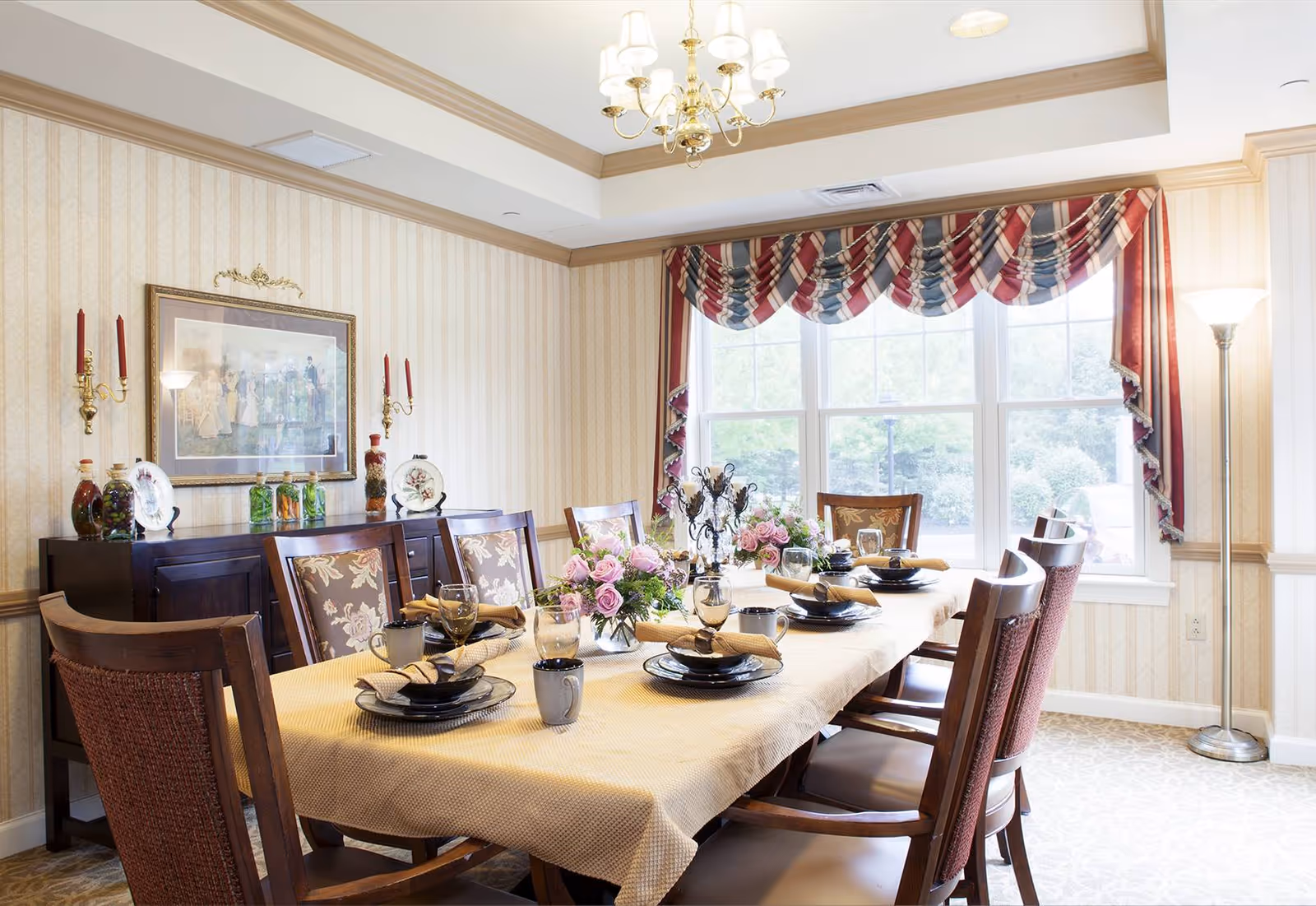 A formal dining room with a long table covered by a beige tablecloth, set with black plates, glasses, mugs, and beige napkins. The table is decorated with pink floral centerpieces. The room has patterned wallpaper, a large window with red, white, and blue valance curtains, a chandelier, and a floor lamp. A dark wooden sideboard against the wall holds decorative items and a framed painting above it.