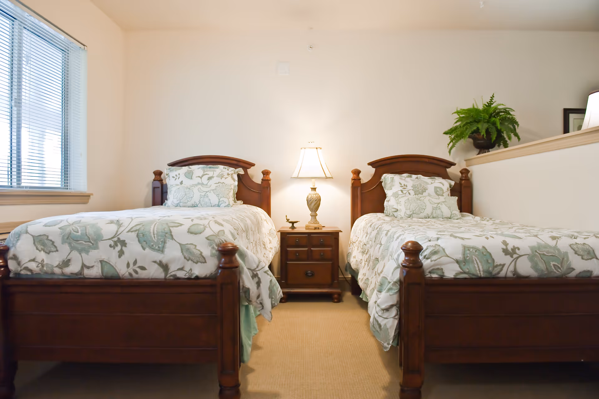 A bedroom with two wooden twin beds featuring floral patterned bedding. Between the beds is a wooden nightstand with a decorative lamp and a small figurine. A window with blinds is on the left wall, and a green potted plant is placed on a half wall to the right.