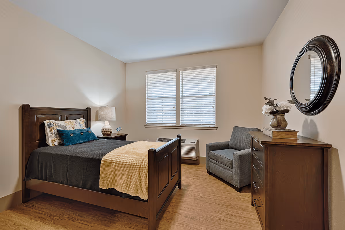 A cozy bedroom with a wooden bed frame, a dark gray bedspread, and a yellow blanket. There are two pillows on the bed, one with a floral pattern and one solid teal. A wooden nightstand with a lamp and a small clock is beside the bed. Across from the bed is a wooden dresser with a vase of white flowers and a round mirror hanging above it. A gray armchair is positioned next to the dresser. The room has two windows with white blinds and wood flooring.