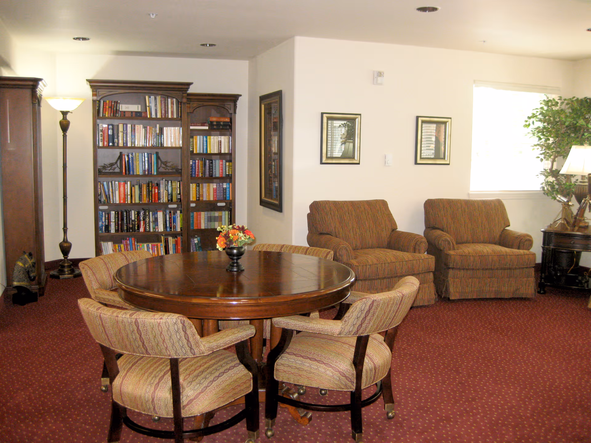 A cozy room with a round wooden table surrounded by four upholstered chairs. Behind the table is a tall wooden bookshelf filled with books. To the right, there are two cushioned armchairs with striped fabric, a side table with a lamp, and a large potted plant near a window. The room has a red carpet with a subtle pattern and light-colored walls with framed artwork.