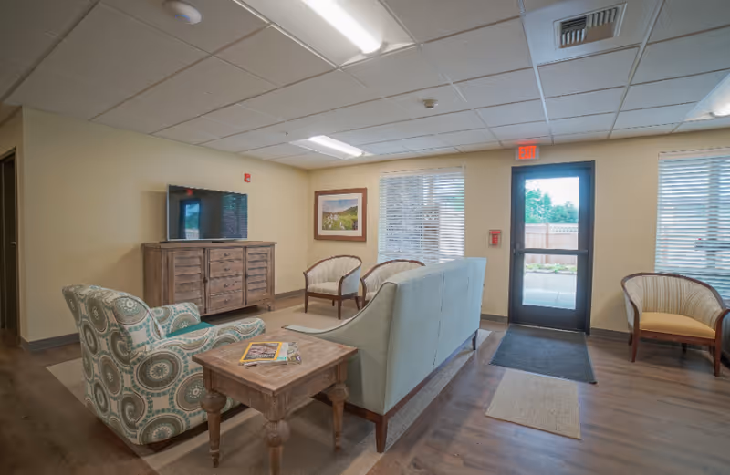 A cozy living room area with a patterned armchair, a wooden side table with magazines, a light blue sofa, and three wooden framed chairs with cushions. A flat-screen TV is mounted on a wooden cabinet against the wall. There are two windows with blinds and a glass door leading outside. The room has wooden flooring and a beige wall color.