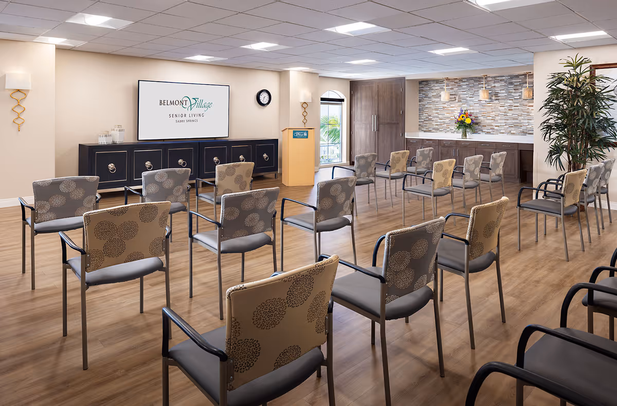 A meeting or presentation room at Belmont Village Senior Living Sabre Springs with rows of patterned chairs facing a large screen displaying the facility's logo. The room has wood flooring, a podium, a clock on the wall, decorative wall sconces, a large plant, and a counter with a flower arrangement against a tiled backsplash.