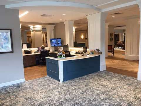 Reception area in a senior living facility with a blue and white front desk, a person working at a computer behind the desk, and a carpeted floor with patterned design. The space has white columns, wood flooring beyond the desk, and seating areas visible in the background.