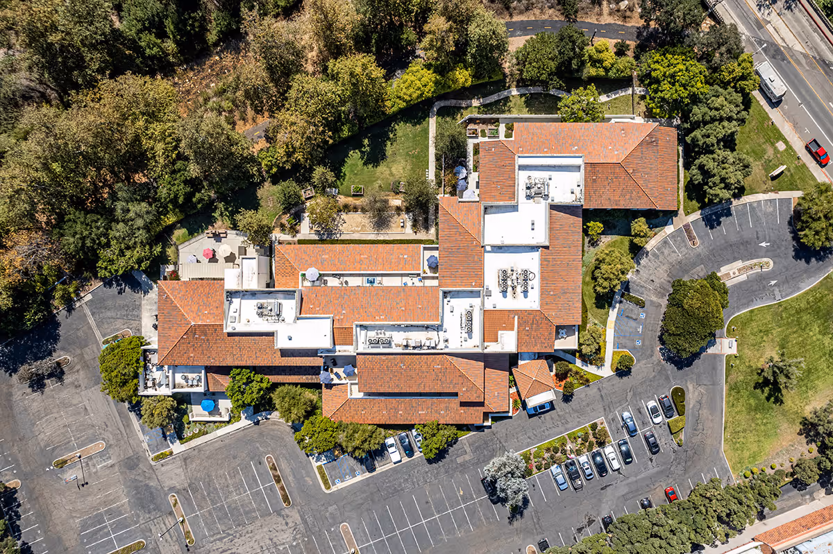 Aerial view of GranVida Senior Living and Memory Care facility showing the building with red-tiled roofs, surrounding parking lots with cars, green landscaped areas, trees, and pathways.