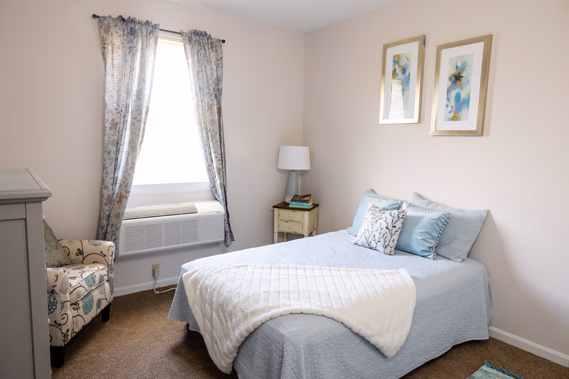 Simple pastel-decor bedroom with a made bed, nightstand and lamp, an armchair and a window with patterned curtains.