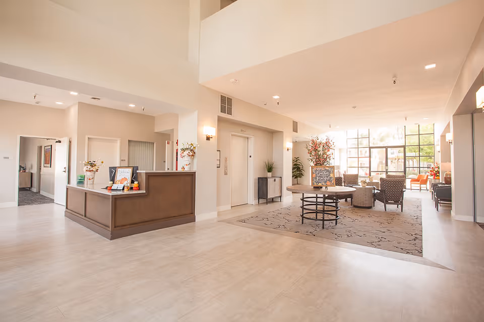 Bright and spacious senior living facility lobby with a reception desk on the left, elevator doors in the center, and a seating area with chairs and a round table on a patterned rug near large windows letting in natural light.
