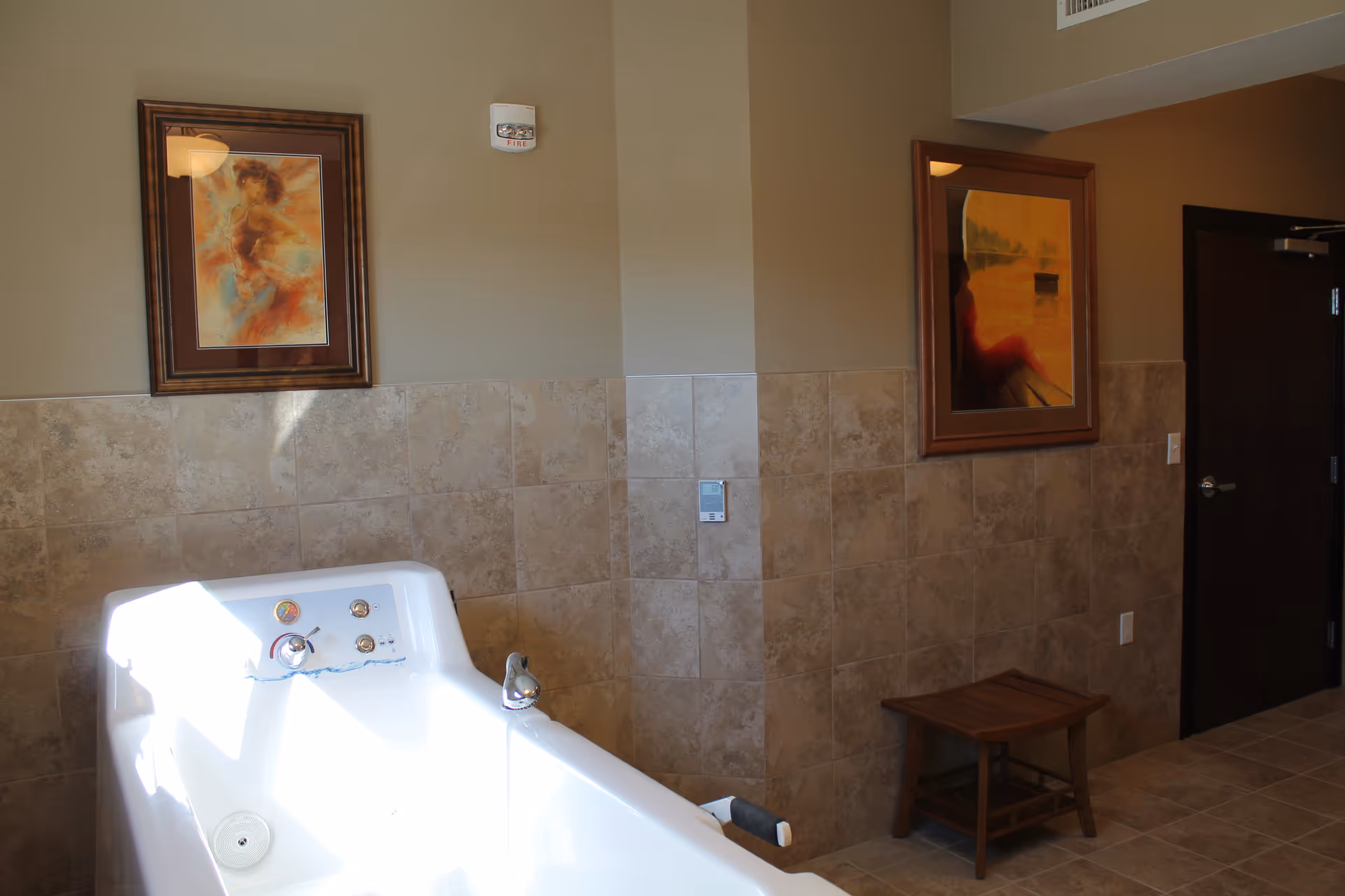 Interior view of a bathroom area with a white bathtub in the foreground. The walls are tiled halfway up with beige tiles and painted beige above. Two framed paintings hang on the walls, one above the bathtub and one near a dark wooden door. A small wooden stool is placed against the wall near the door.