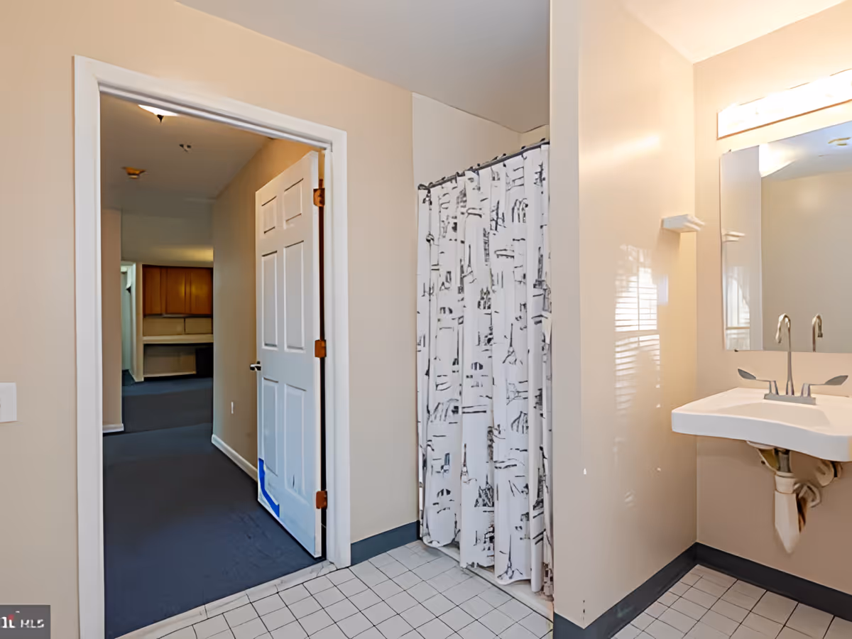Interior view of a bathroom in the Golden Arms II facility showing a white sink with a mirror above it, a shower area with a white curtain featuring black patterns, beige walls, and a white door leading to a carpeted hallway.