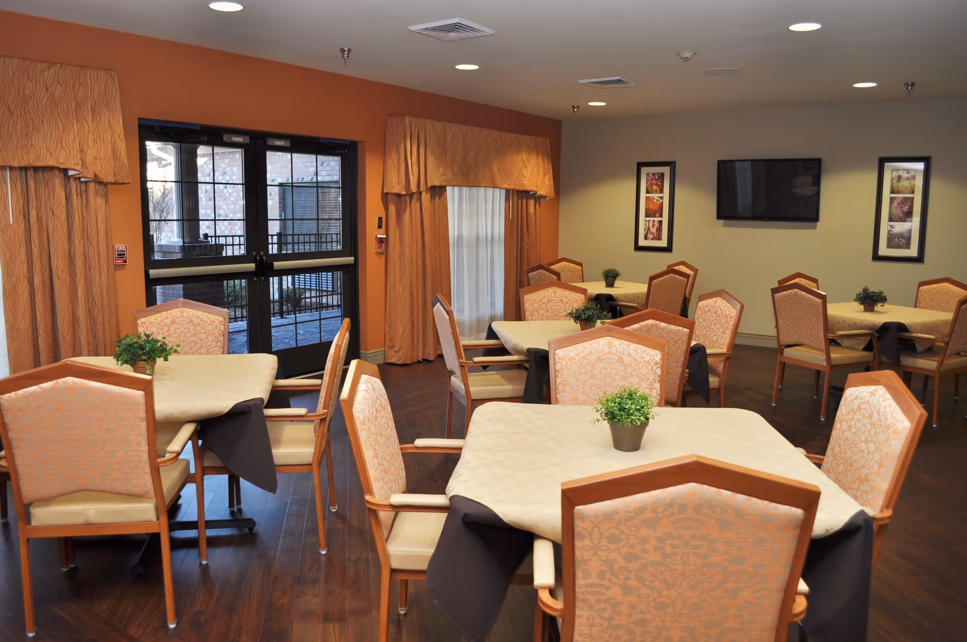 A dining room with several tables covered with beige tablecloths and black underlays. Each table has four chairs with patterned upholstery and a small green plant centerpiece. The room has wooden flooring, orange curtains, and a glass door leading outside. On the far wall, there is a flat-screen TV mounted between two framed pictures.