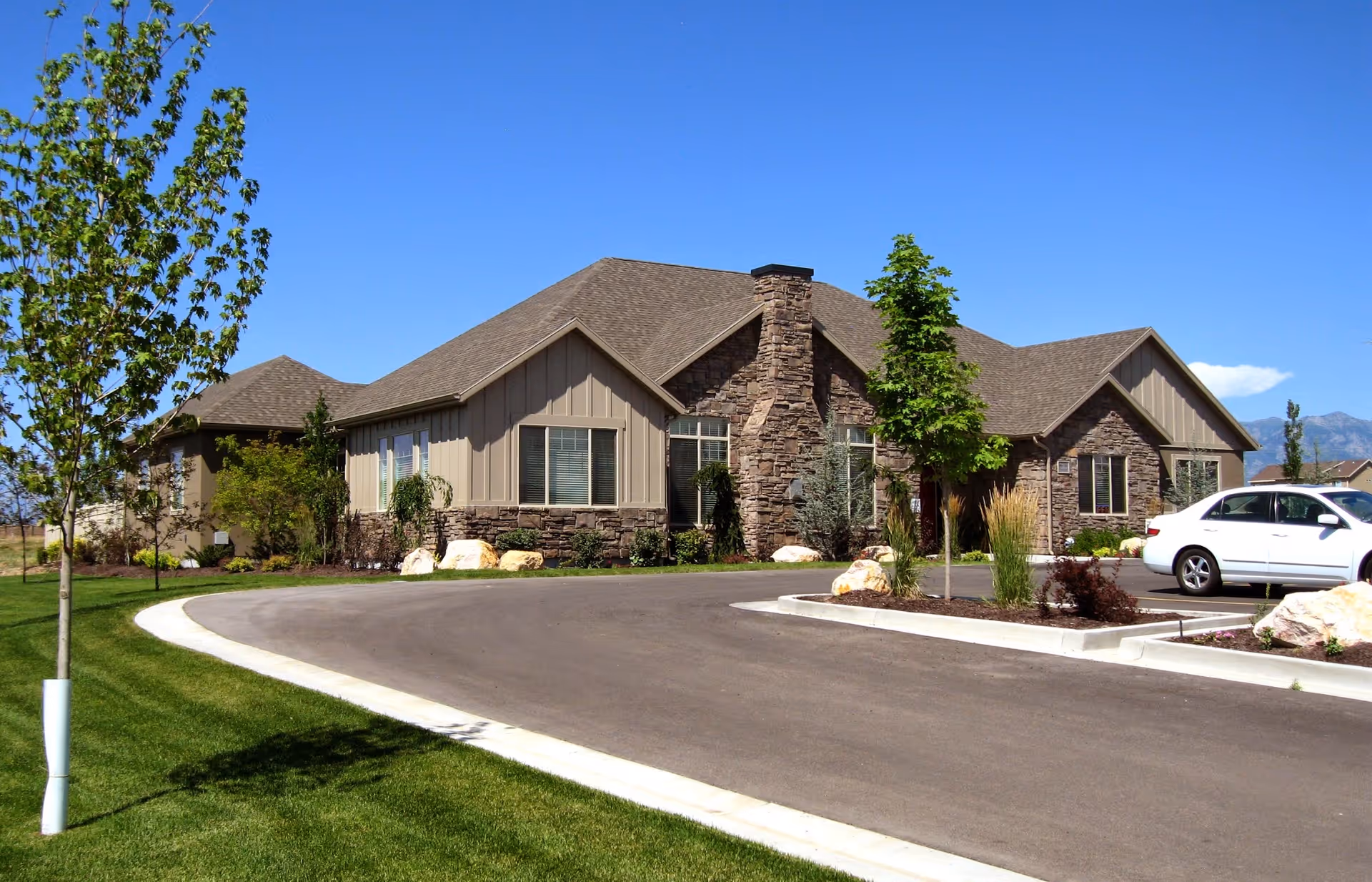 Exterior view of a single-story building with stone and siding facade, surrounded by landscaped greenery, a curved driveway, and a parked white car under a clear blue sky.