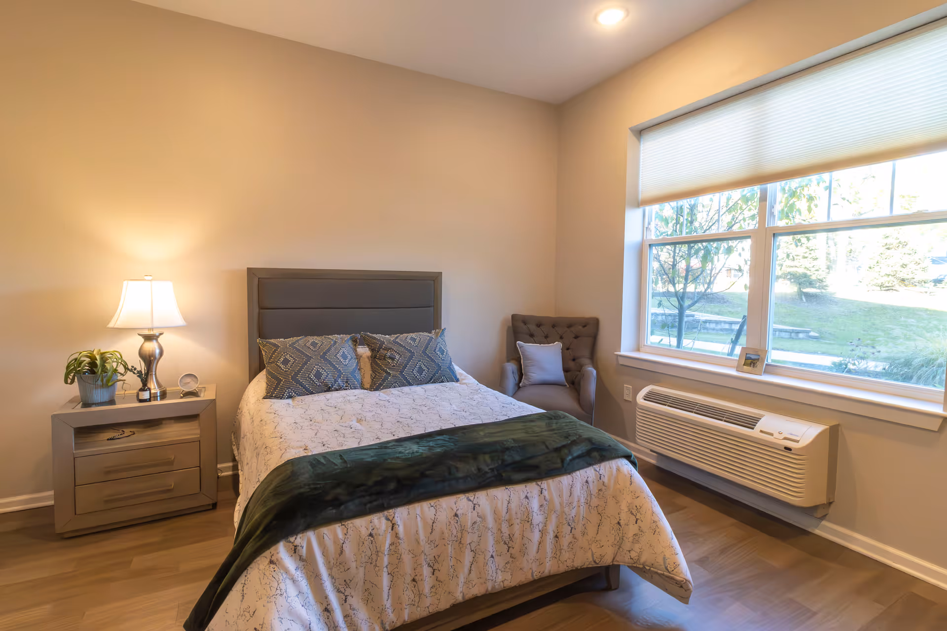 A cozy bedroom with a neatly made bed featuring patterned pillows and a green throw blanket. To the left of the bed is a nightstand with a lamp, a small plant, and a clock. A cushioned armchair with a pillow is positioned in the corner near a large window with a view of greenery outside. Below the window is a wall-mounted air conditioning unit.