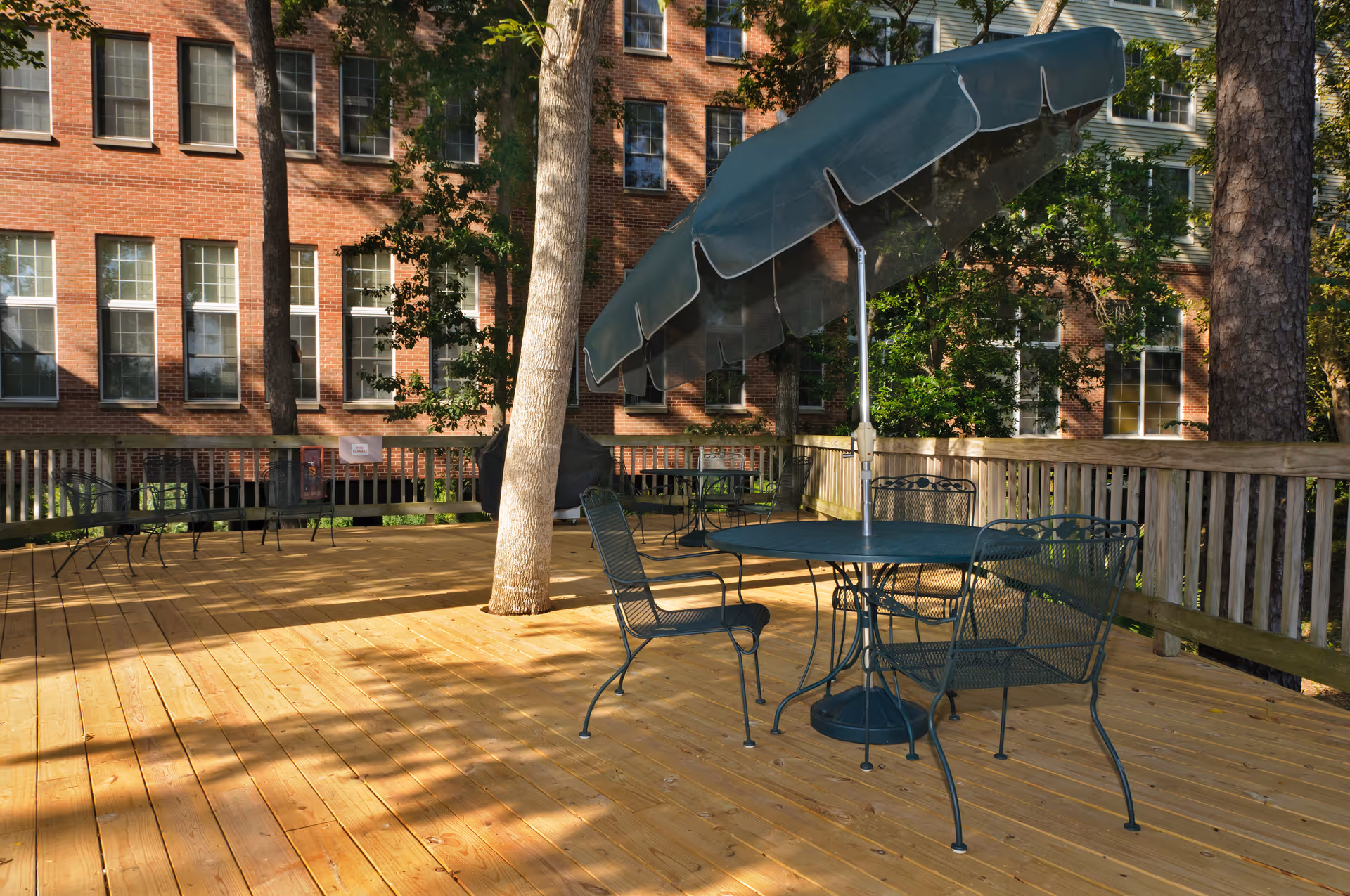 Outdoor wooden deck area with metal patio tables and chairs, one table with a large green umbrella. Trees are growing through the deck, and a brick building with multiple windows is visible in the background.