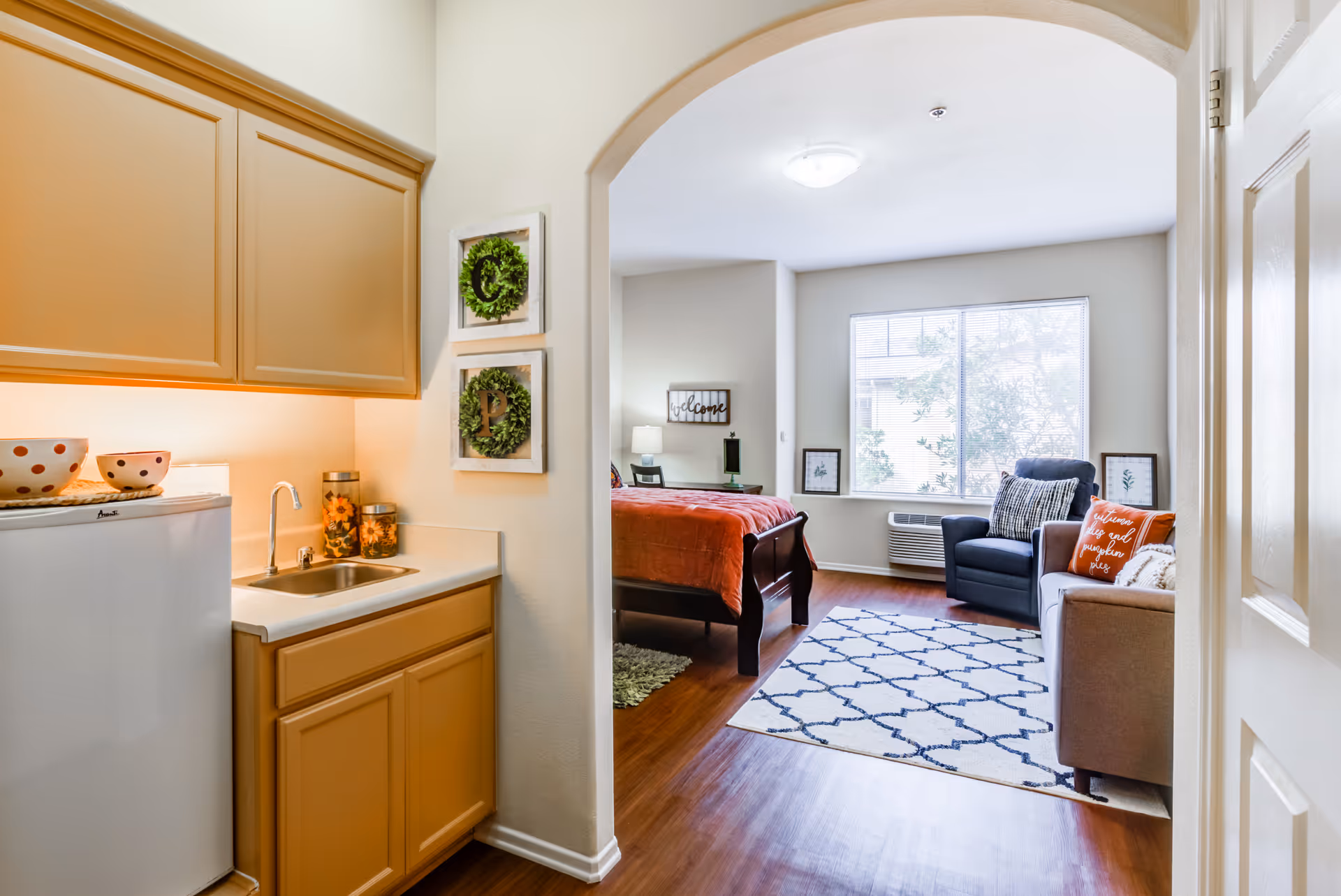 View of a small kitchenette with a sink, countertop, and cabinets on the left, leading through an arched doorway into a living area with a bed, armchair, sofa, and a large window letting in natural light. The living area has a patterned rug on the wooden floor and decorative pillows on the sofa.