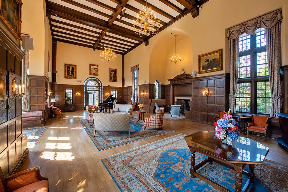 Spacious and elegant living room with high wooden beamed ceilings, large windows with drapes, chandeliers, wood-paneled walls, a grand piano, multiple seating areas with sofas and armchairs, decorative rugs on a wooden floor, and a fireplace.
