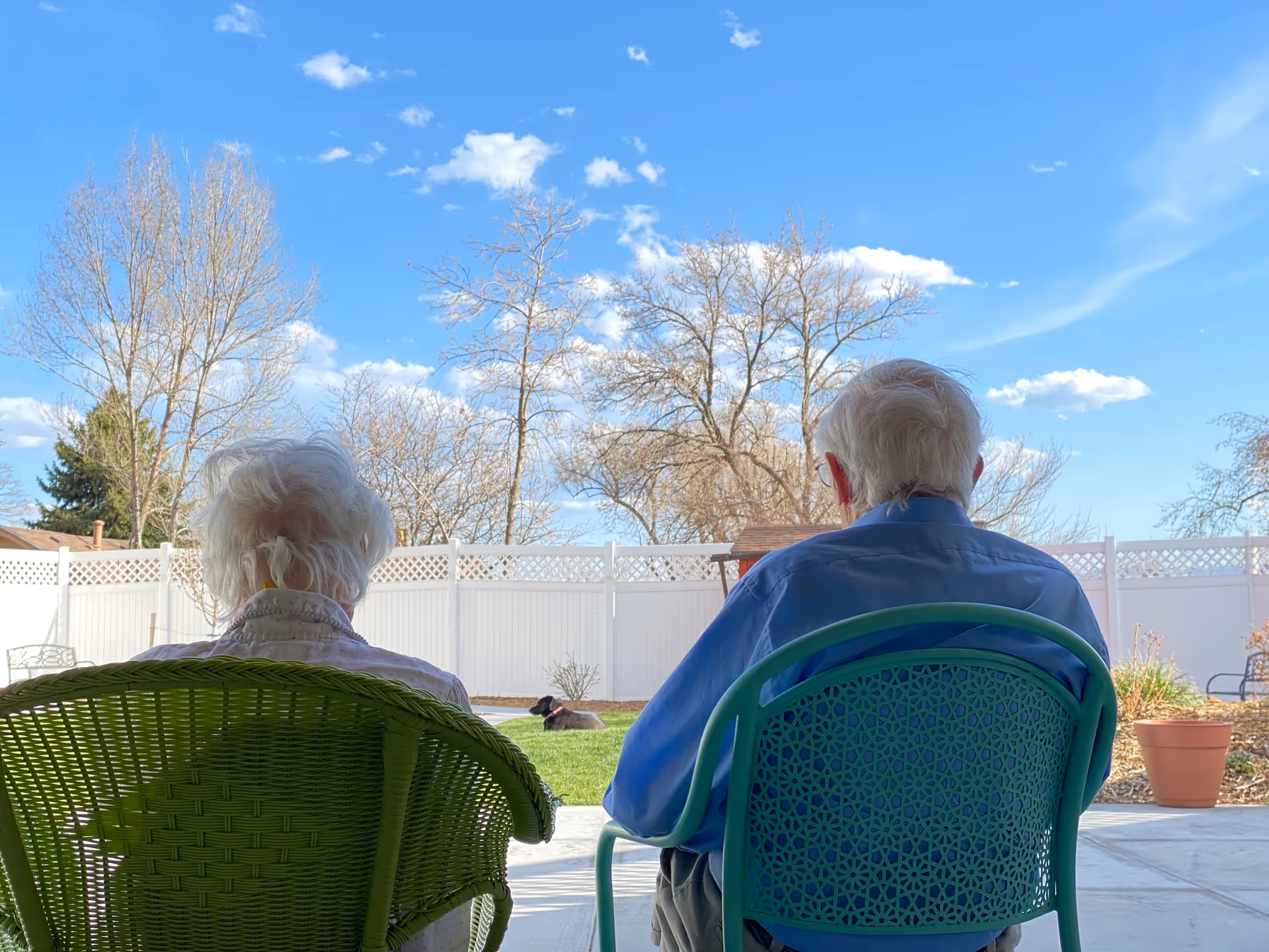 Two elderly individuals sitting outdoors on green chairs facing away, looking at a fenced backyard with leafless trees and a dog lying on the grass under a blue sky with scattered clouds.