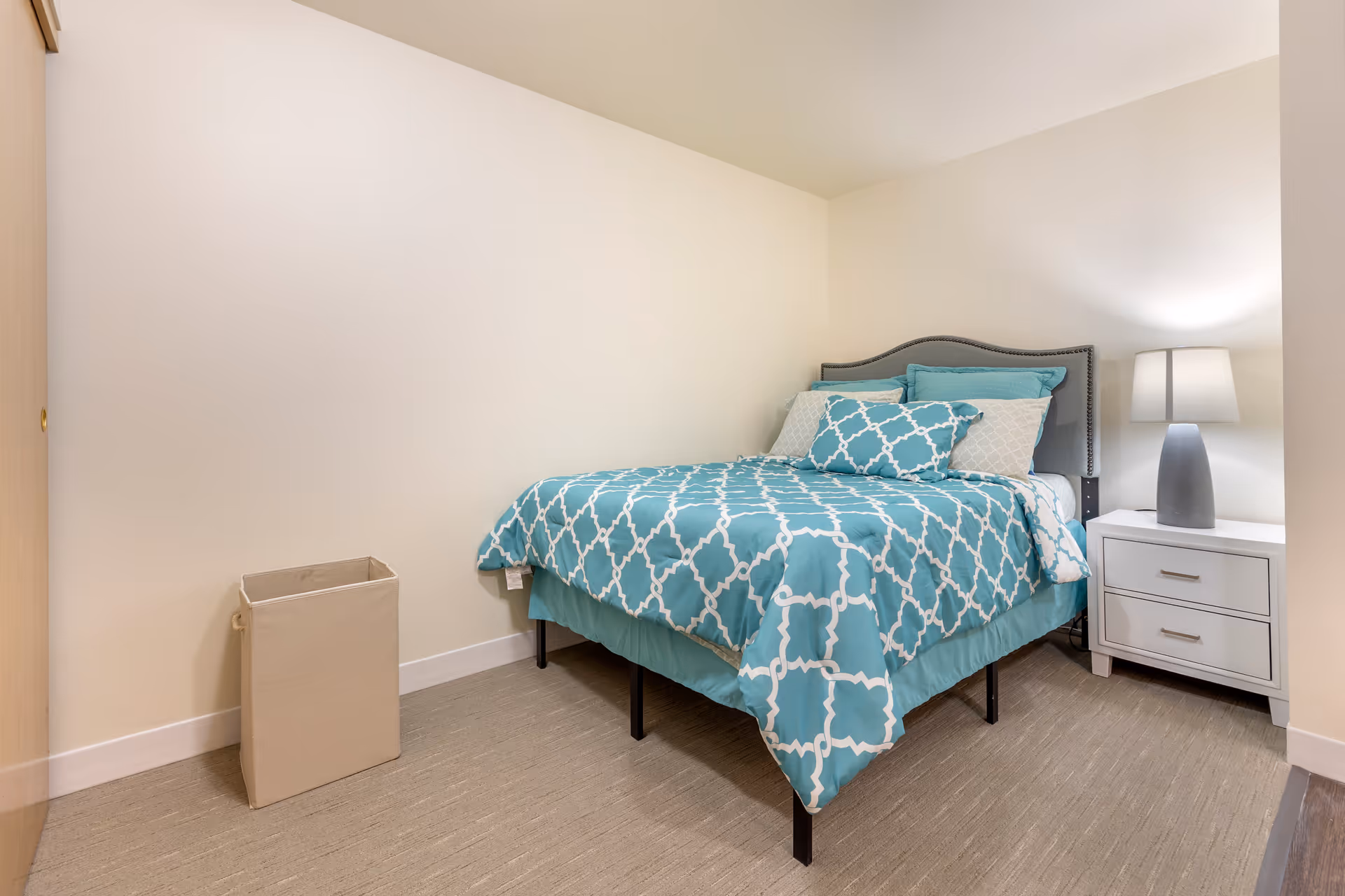 Small bedroom with a teal patterned bed, a white nightstand and lamp, and a linen hamper against a cream wall.