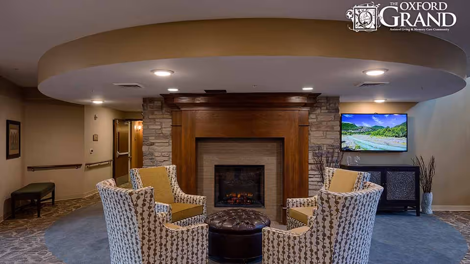 Cozy sitting area with four patterned armchairs arranged around a fireplace and a wall-mounted TV in a common room.
