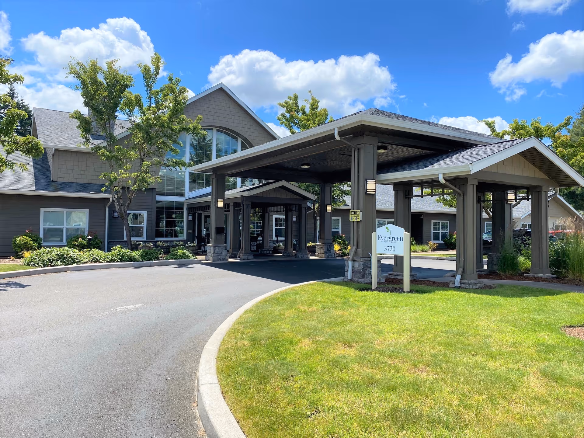 Exterior front entrance of Evergreen Memory Care showing the covered porte-cochere, building facade, and lawn under a blue sky.