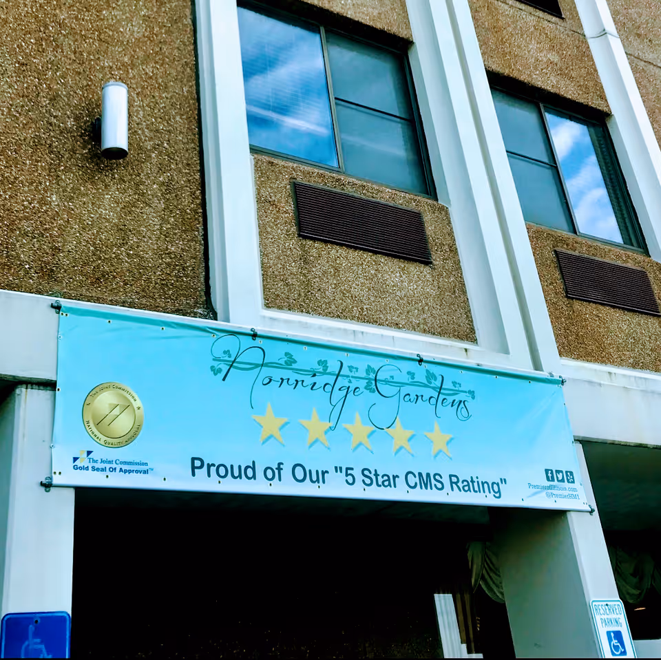 Exterior view of a building with a banner that reads 'Norridge Gardens Proud of Our 5 Star CMS Rating' along with five gold stars and a gold seal of approval from The Joint Commission. The building has two windows and a reserved parking sign for handicapped parking is visible.