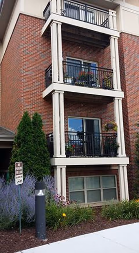 Exterior view of a brick building with three balconies featuring black railings and potted plants. There are shrubs and flowers planted along the base of the building near a sidewalk.