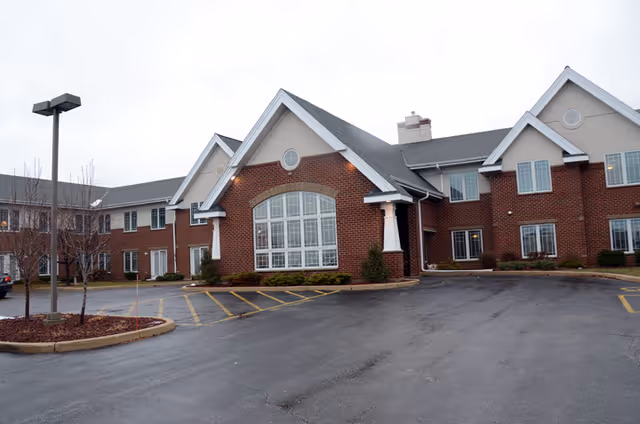 Front exterior of a two-story red brick assisted living building with a large arched window and a wet parking lot under an overcast sky.