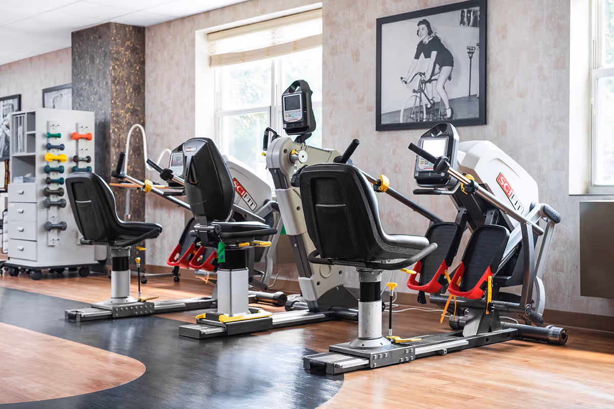 A rehabilitation gym room with three seated exercise machines designed for physical therapy. The room has wooden flooring with a black mat area, a rack of colorful dumbbells, and a large window letting in natural light. A black and white photo of a woman on a bicycle is hung on the wall.