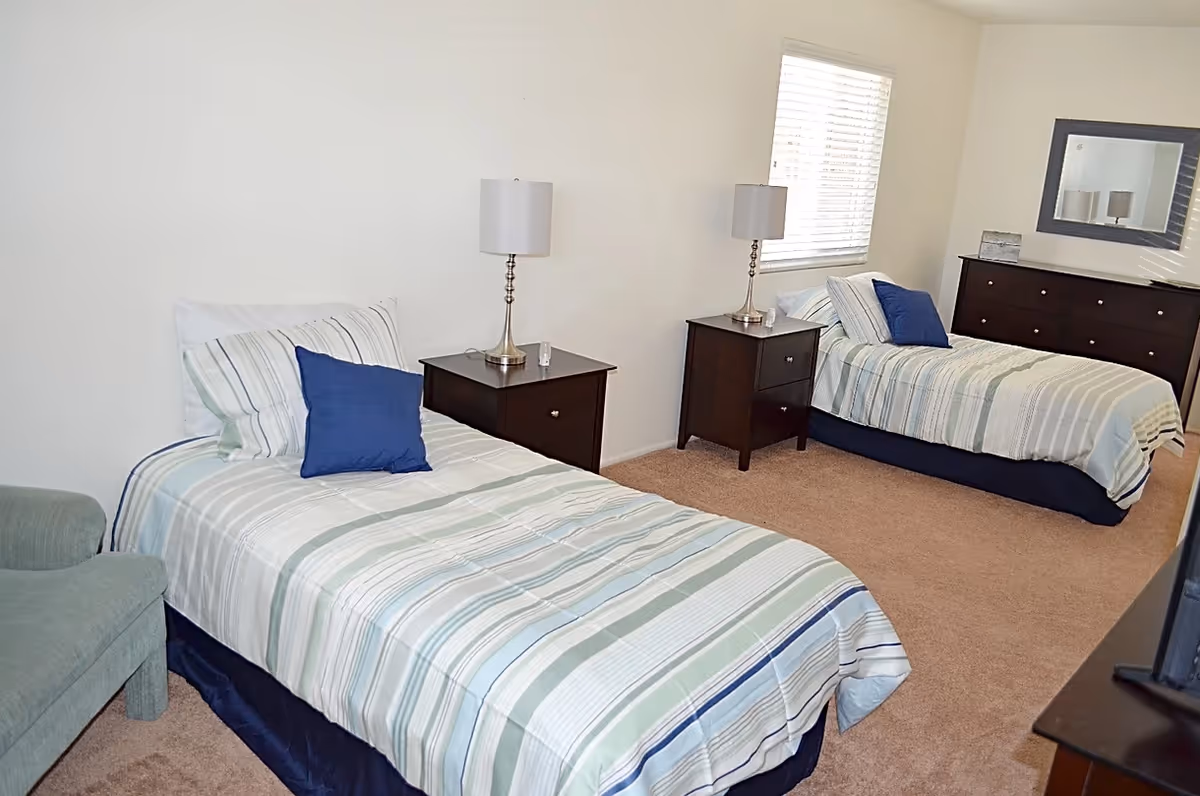 A bright bedroom with two twin beds dressed in striped bedding, nightstands with lamps, and a dresser with a mirror.