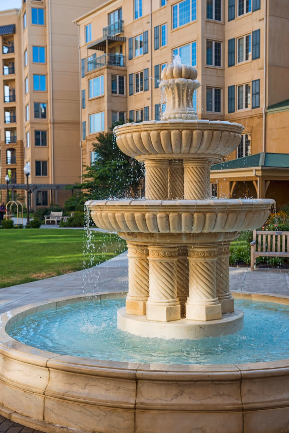 A multi-tiered stone water fountain with water cascading down into a circular basin, set in an outdoor courtyard area with green grass, benches, and a multi-story beige building with many windows in the background.