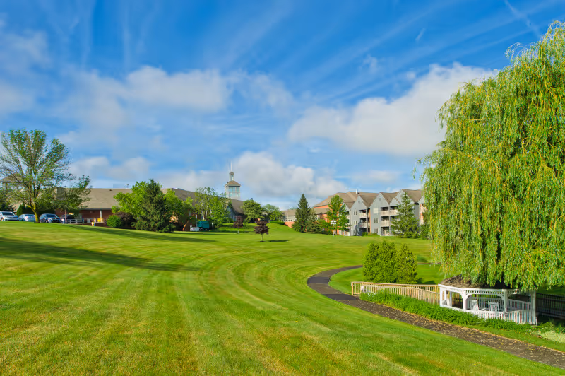 A large, well-maintained grassy lawn with a paved walking path curving through it. There are several trees, including a large weeping willow on the right side near a white gazebo. In the background, there are multi-story residential buildings and a building with a cupola under a partly cloudy blue sky.