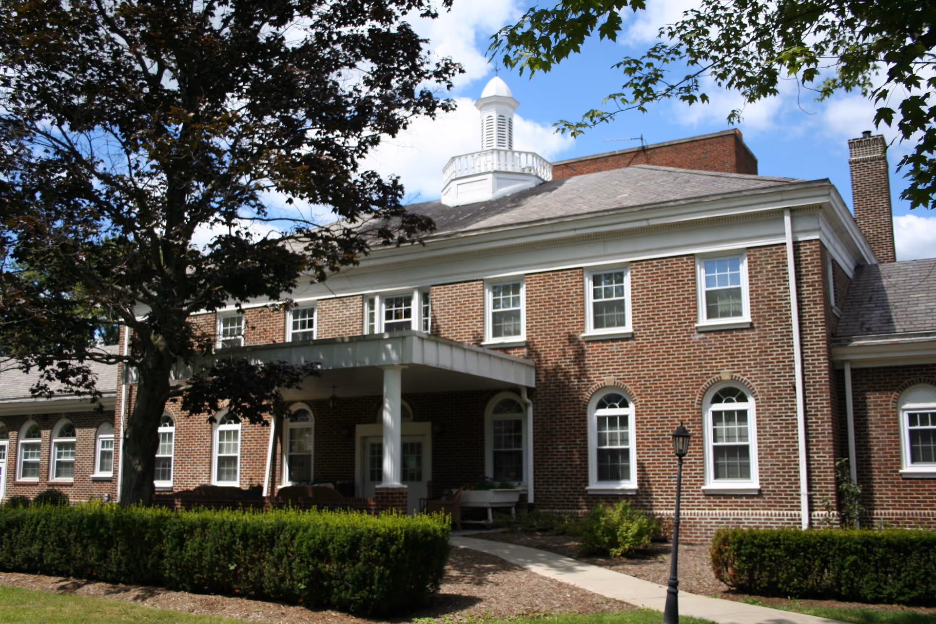 Exterior view of a large brick building with white-trimmed windows and a covered entrance. The building is surrounded by green bushes and trees under a partly cloudy blue sky.