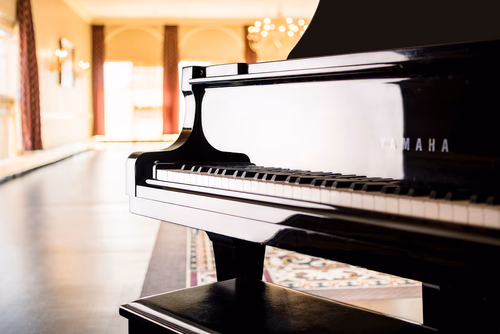 Close-up view of a black Yamaha grand piano with a matching bench in a spacious, well-lit room featuring large windows and chandeliers in the background.