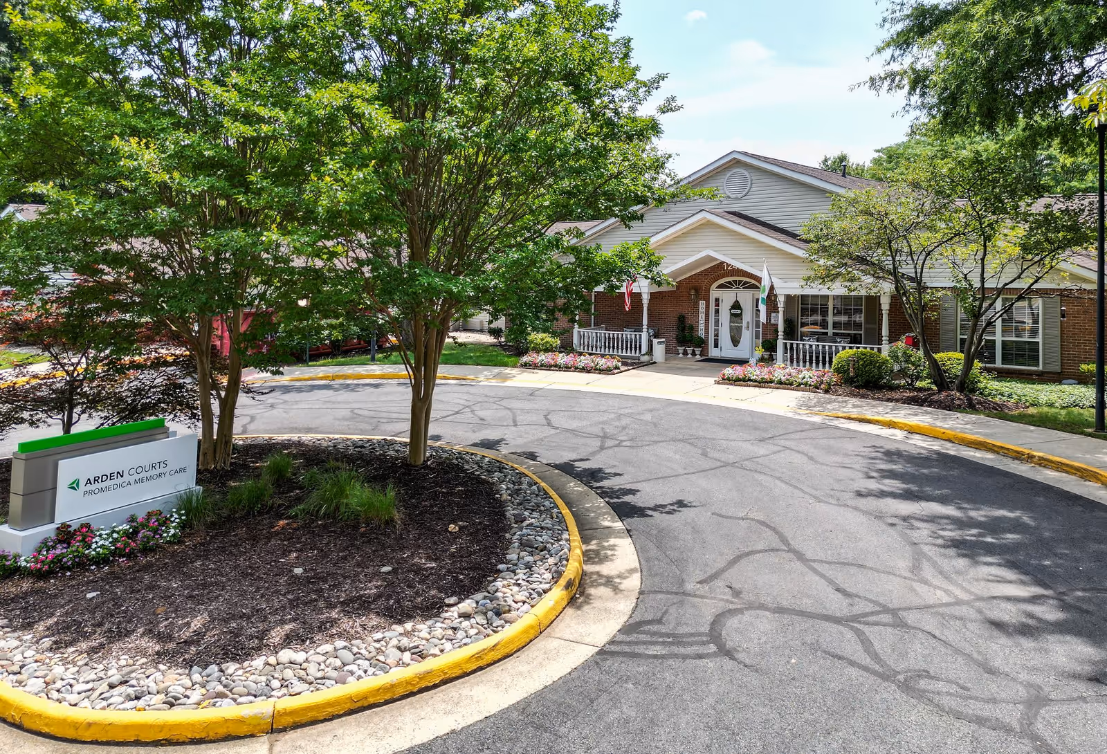 Front exterior view of Arden Courts - ProMedica Memory Care Community building with a circular driveway, trees, and landscaping including flowers and bushes. A sign with the facility name is visible near the driveway.