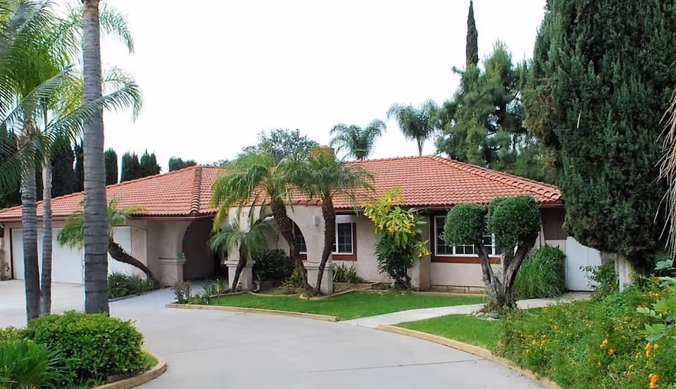 Single-story building with a red-tiled roof, arched entrance, palm trees and landscaped driveway in front.