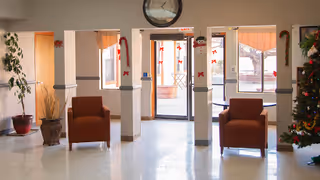 A bright and clean common area with two brown armchairs placed near white columns decorated with red bows. There is a large clock above a glass door leading outside, and a Christmas tree decorated with ornaments is visible on the right side. Potted plants and a window with curtains are also seen in the background.