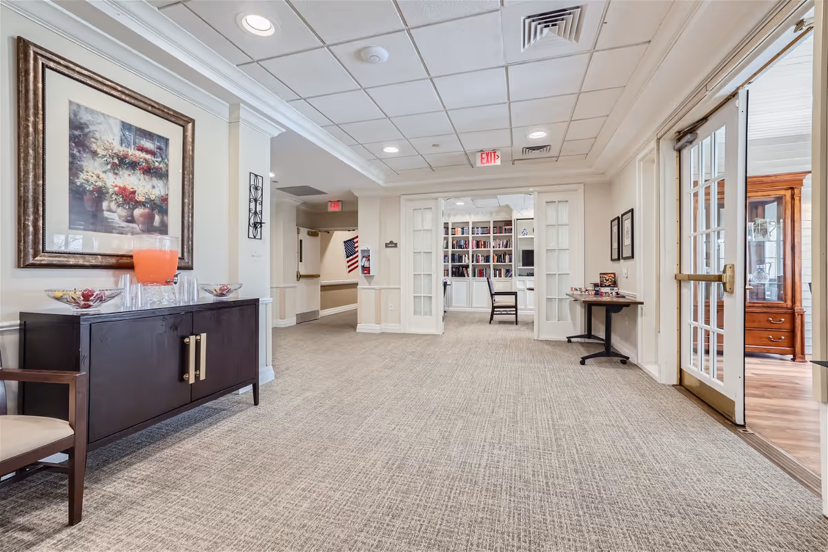 Interior hallway of Bear Creek Assisted & Senior Living featuring beige carpet, cream-colored walls, and a ceiling with recessed lighting. On the left side, there is a dark wooden cabinet with a large framed floral painting above it and a pitcher of orange drink with cups. At the end of the hallway, double glass doors open into a room with bookshelves and a chair. On the right, there is a glass door leading to another room with wooden furniture visible.