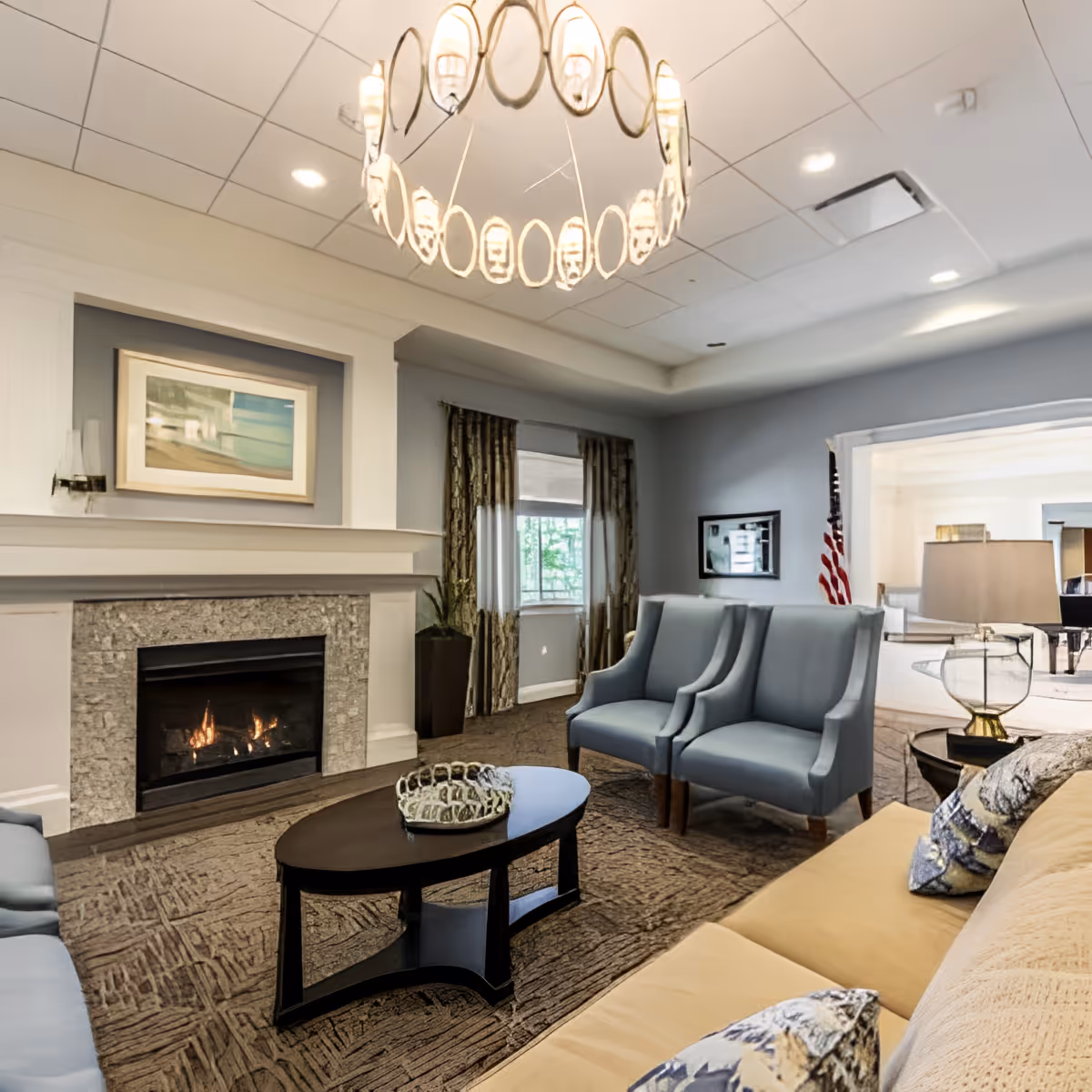A cozy living room with a lit fireplace, a modern circular chandelier, a dark wooden coffee table, two blue armchairs, a beige sofa with patterned pillows, and large windows with curtains. The room has a neutral color palette with light gray walls and carpeted floor.