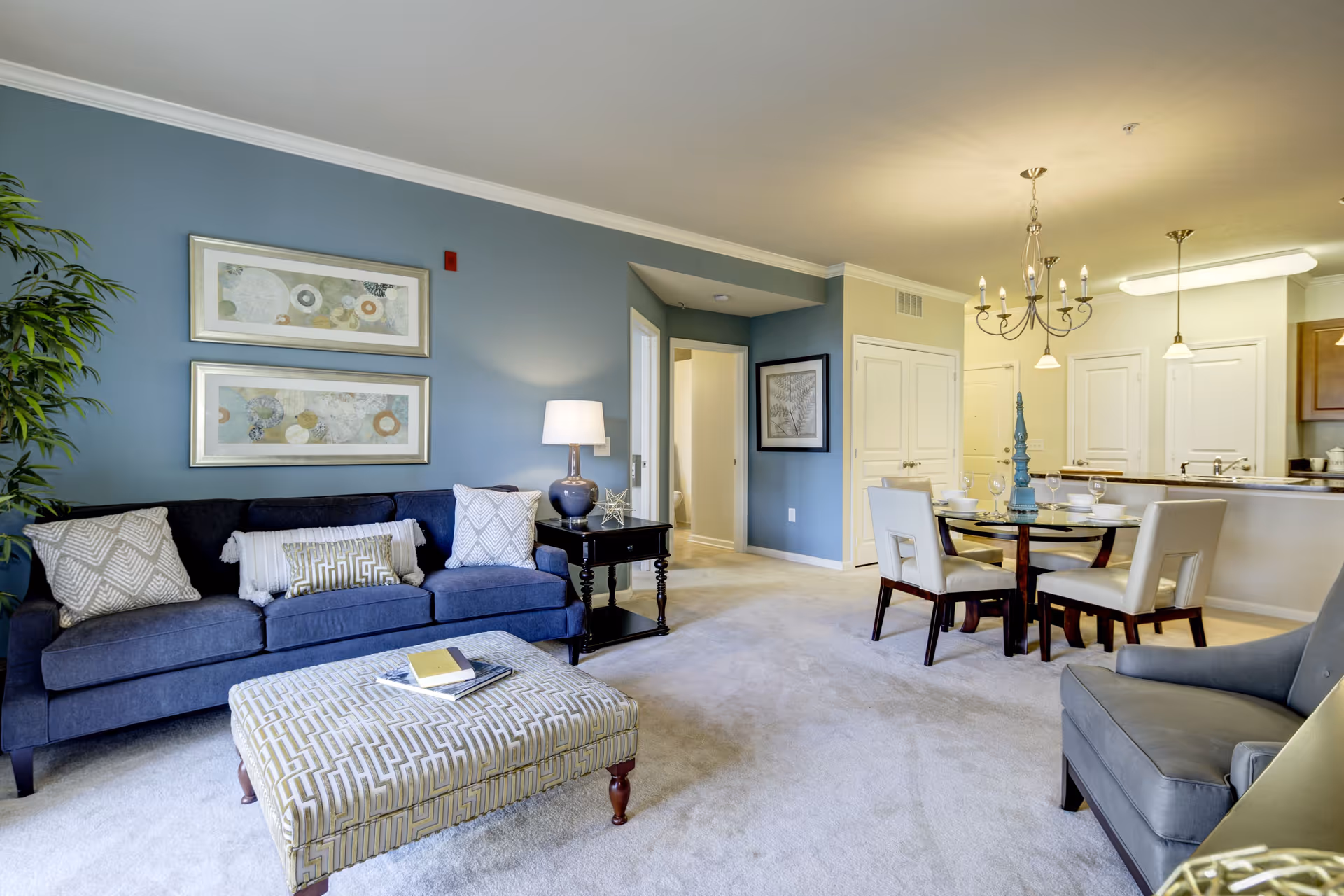 A bright and spacious living room area with a blue sofa adorned with patterned pillows, a matching upholstered ottoman with books on top, a side table with a lamp, and a gray armchair. The room features light carpet flooring, blue and beige walls, framed artwork, and a dining area with a round glass table and four white chairs. The kitchen is visible in the background with wooden cabinets and a breakfast bar.