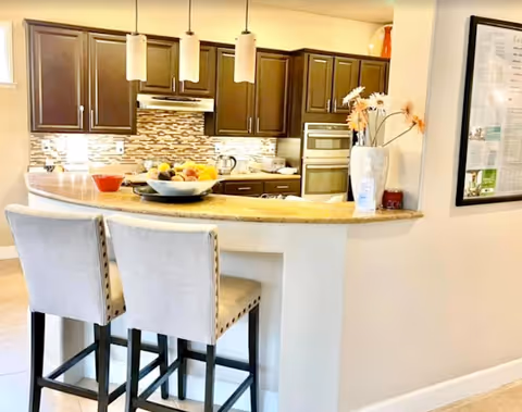 A modern kitchen with dark wooden cabinets, a tiled backsplash, and stainless steel appliances including a built-in oven and range hood. There is a curved countertop with two upholstered bar stools, a bowl of fruit, and a vase with flowers on the counter. Three pendant lights hang above the counter.
