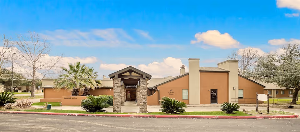 Front exterior of a single-story tan assisted living facility with a stone entrance, palm trees, and landscaped grounds under a blue sky.