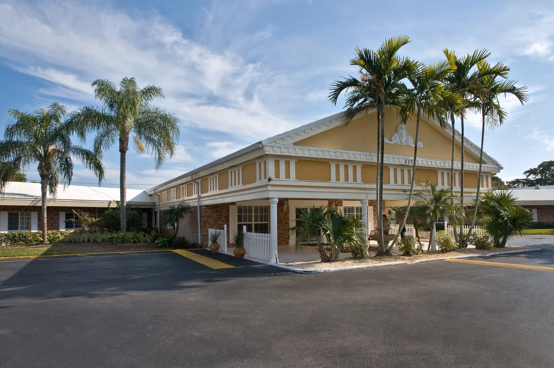 Exterior view of Darcy Hall of Life Care building with a yellow and white facade, surrounded by palm trees and landscaping under a partly cloudy sky.