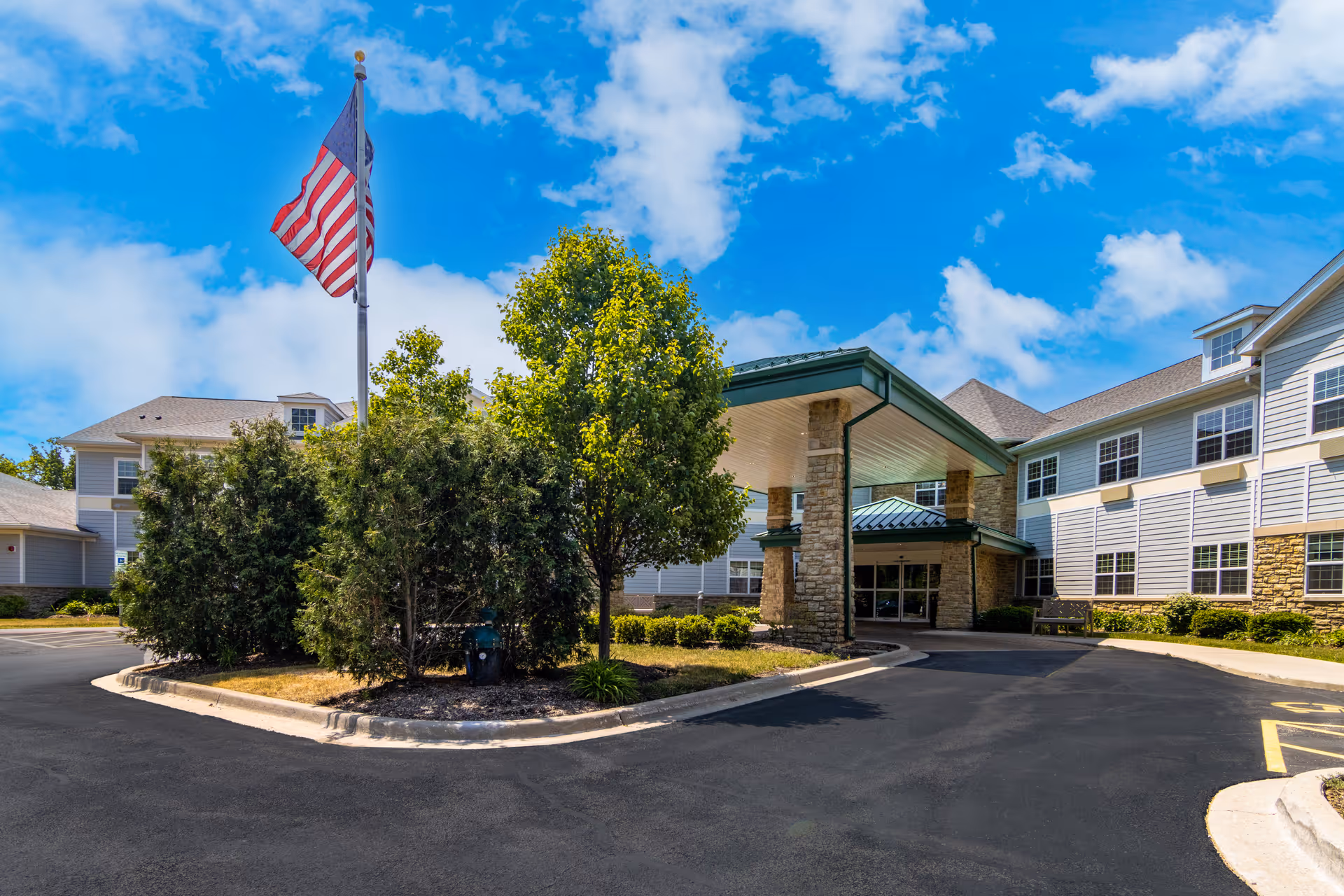 Exterior view of New Perspective Senior Living | Long Grove facility showing the entrance with a covered driveway, surrounded by trees and bushes, an American flag on a flagpole, and a bright blue sky with some clouds.