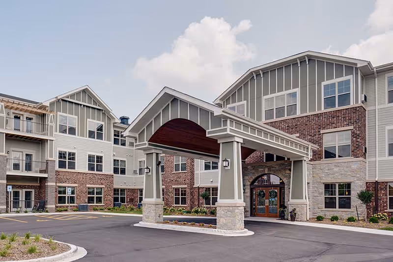 Exterior view of Aspire Oshkosh senior living facility showing a large covered entrance with stone pillars, brick and siding facade, multiple windows, and a paved driveway under a partly cloudy sky.
