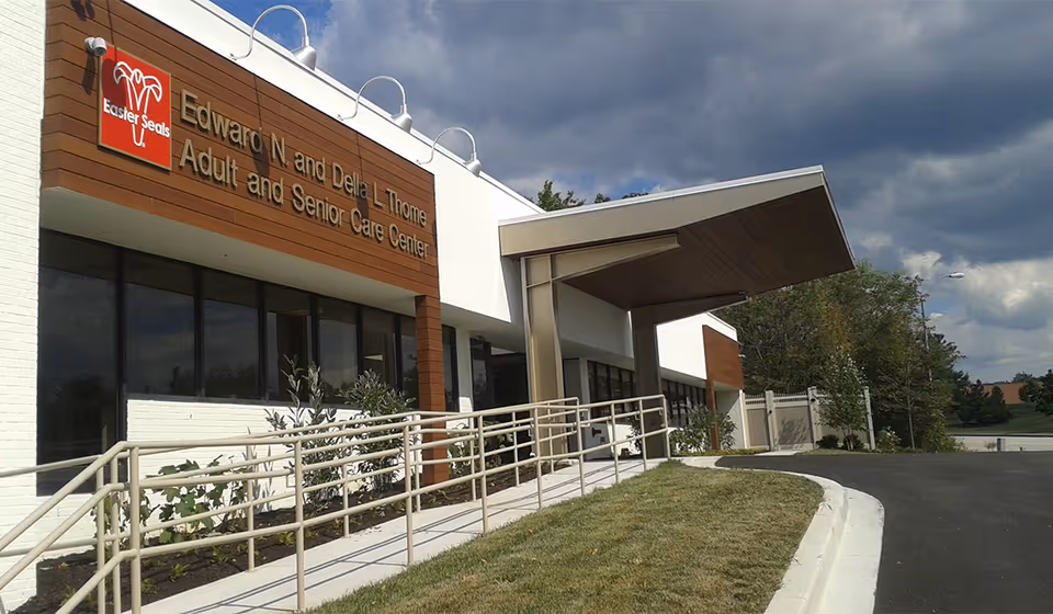 Exterior view of the Edward N. and Della L. Thome Adult and Senior Care Center building with a ramp leading to the entrance. The building has a modern design with white walls, wooden paneling, and large windows under a cloudy sky.
