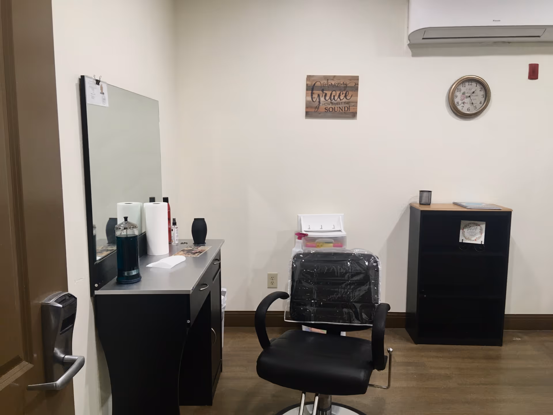 Interior grooming/salon room with a black styling chair in front of a mirror, a counter with supplies, and a small bookshelf against a plain wall.