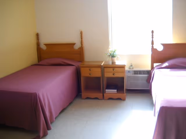 A simple bedroom with two single beds covered in purple bedspreads, separated by two wooden nightstands. A small plant and a decorative item are placed on the nightstands. There is a window with bright sunlight coming through, and an air conditioning unit below the window.